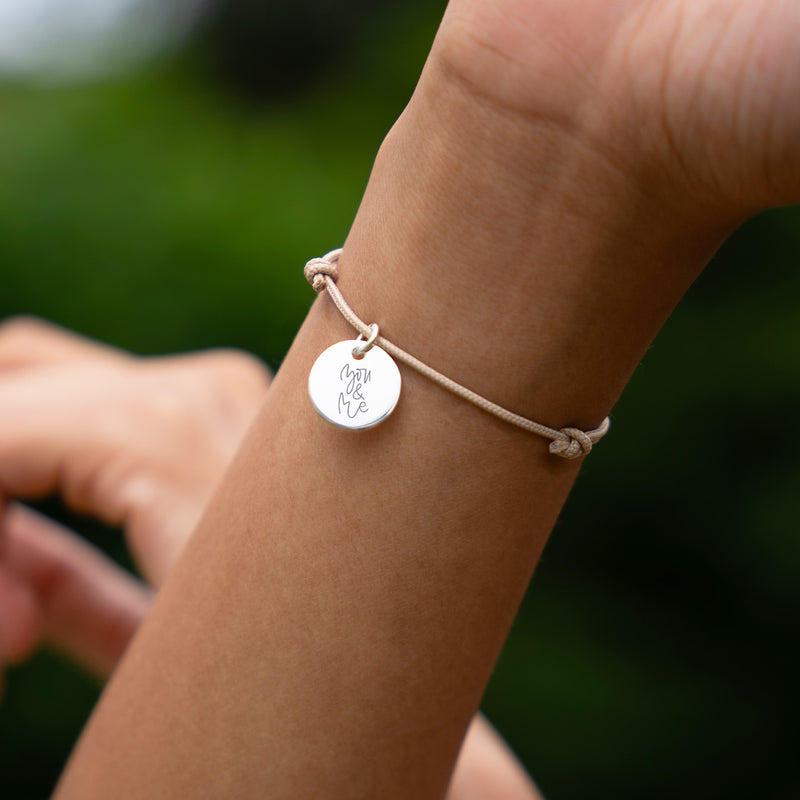 Bracelet personnalisé en argent, avec une médaille de 15mm, sur un cordon de couleur beige.
