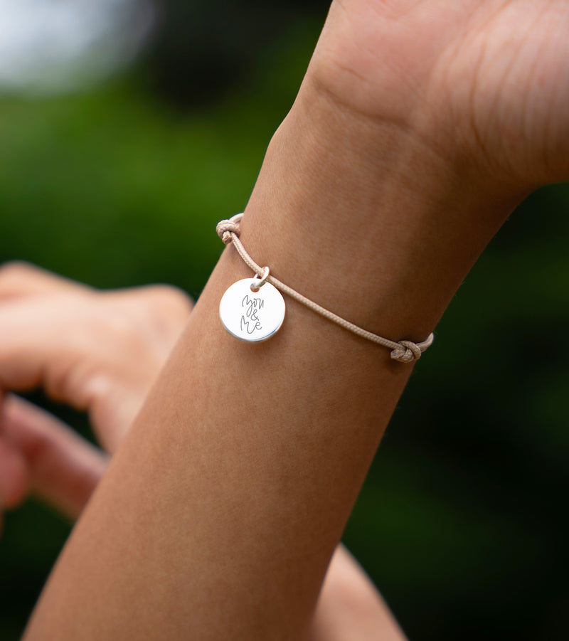 Bracelet personnalisé en argent, avec une médaille de 15mm, sur un cordon de couleur beige.