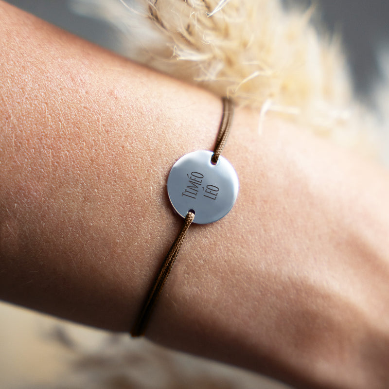 Un bracelet argenté orné d'une médaille de 15mm et de deux trous traversée par un cordon en marron clair, unique en son genre.