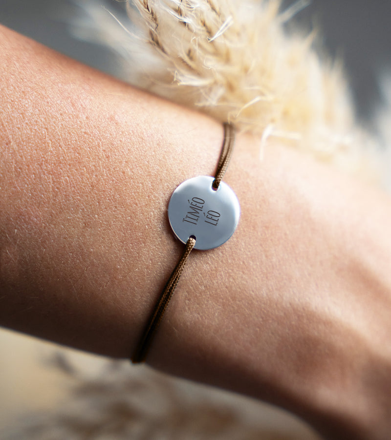 Un bracelet argenté orné d'une médaille de 15mm et de deux trous traversée par un cordon en marron clair, unique en son genre.