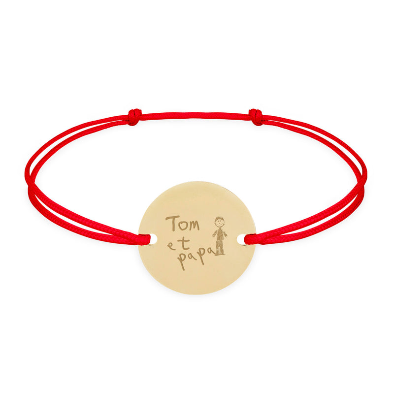 Un bracelet personnalisé, doté d'une médaille plaquée or de 15mm, accrochée par deux trous, sur cordon rouge.