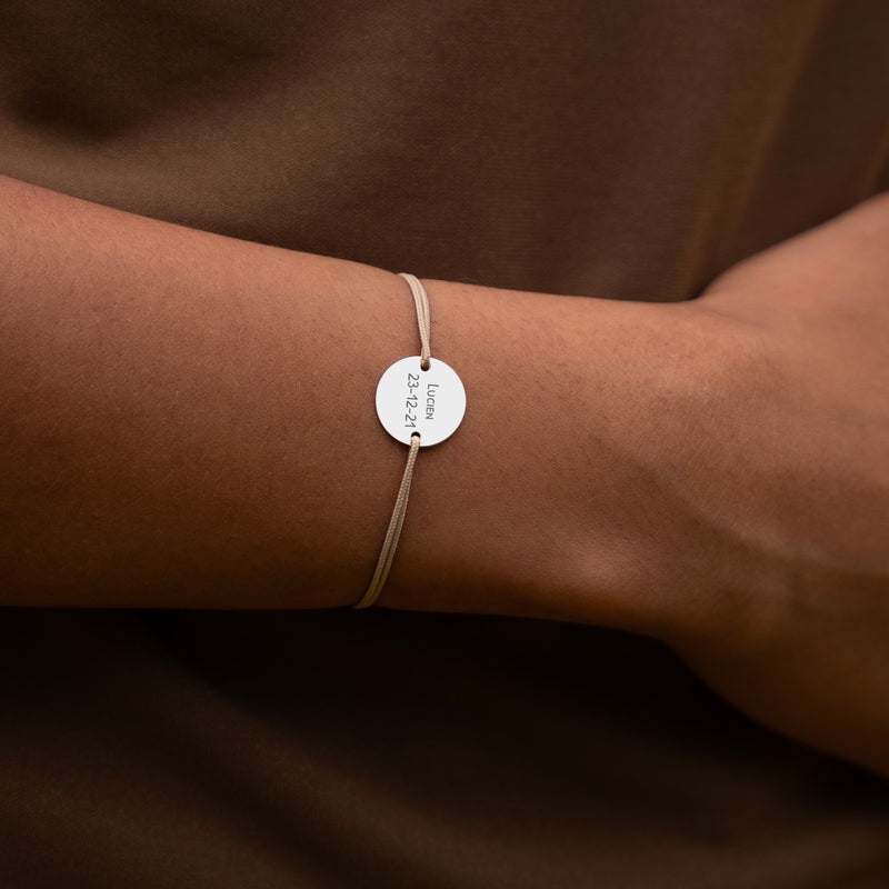 Une médaille personnalisée de 15mm montée sur un bracelet argenté, accompagnée de deux trous pour un cordon beige.