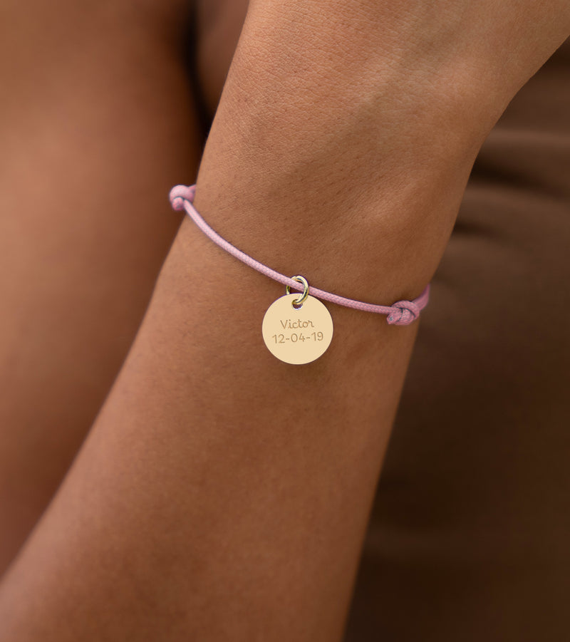 Un bijou composé d'une médaille plaquée or de 15mm suspendue à un cordon rose est illustré.