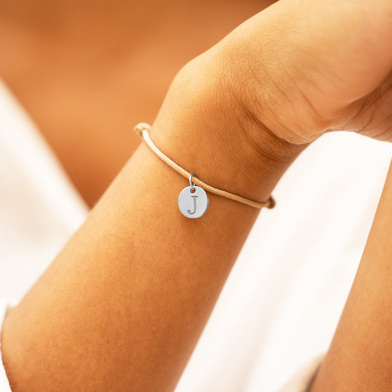 Médaille argentée de 10mm suspendue sur un bracelet en cordon beige.