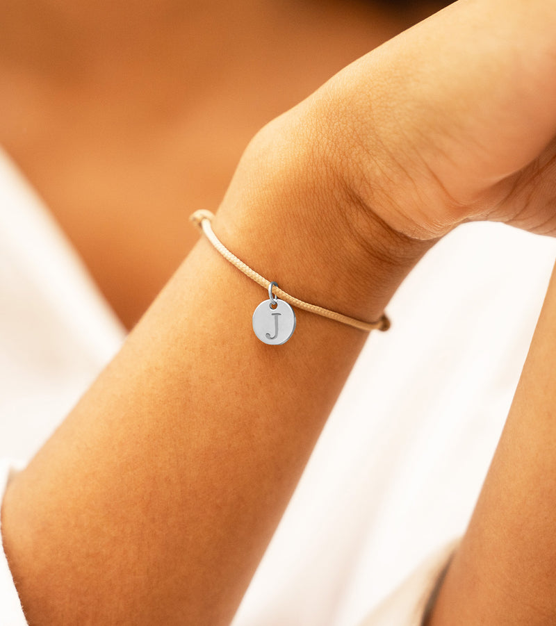 Médaille argentée de 10mm suspendue sur un bracelet en cordon beige.