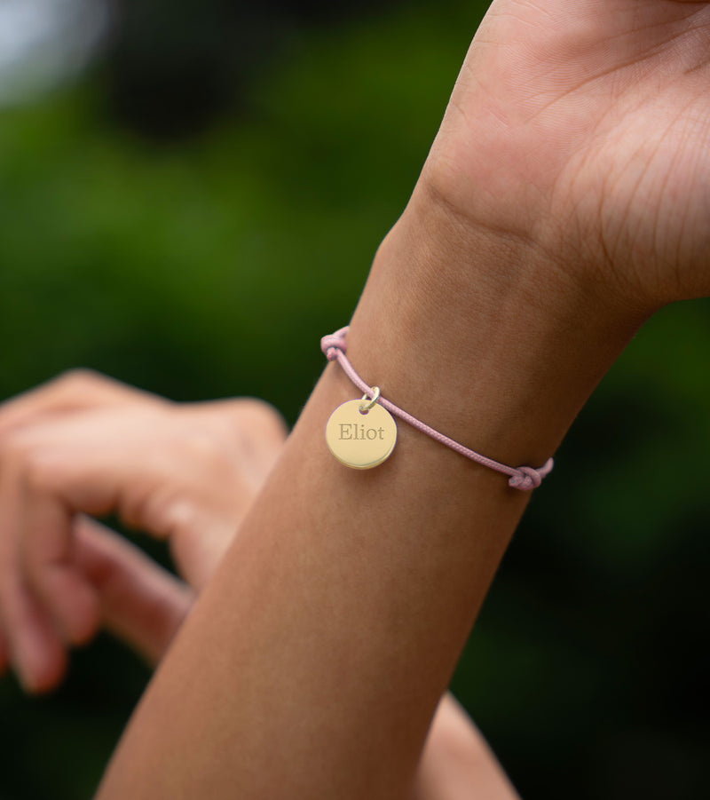 La photo montre un bracelet personnalisé avec une médaille de 15mm en plaqué or, attachée à un cordon rose.