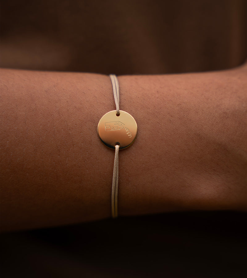 Un bracelet personnalisé, orné d'une médaille plaquée or de 15mm et de deux trous, complété par un cordon beige.