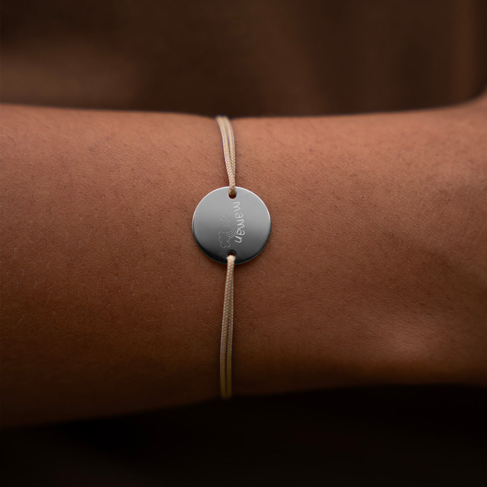Un bracelet argenté, mettant en valeur une médaille de 15mm personnalisable, avec deux trous pour un cordon beige. 