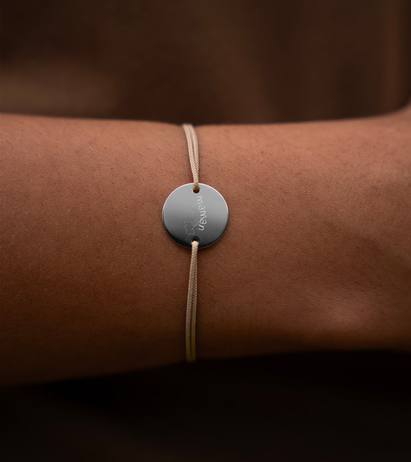 Un bracelet argenté, mettant en valeur une médaille de 15mm personnalisable, avec deux trous pour un cordon beige. 