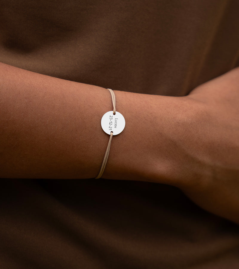 Une médaille personnalisée de 15mm montée sur un bracelet argenté, accompagnée de deux trous pour un cordon beige.