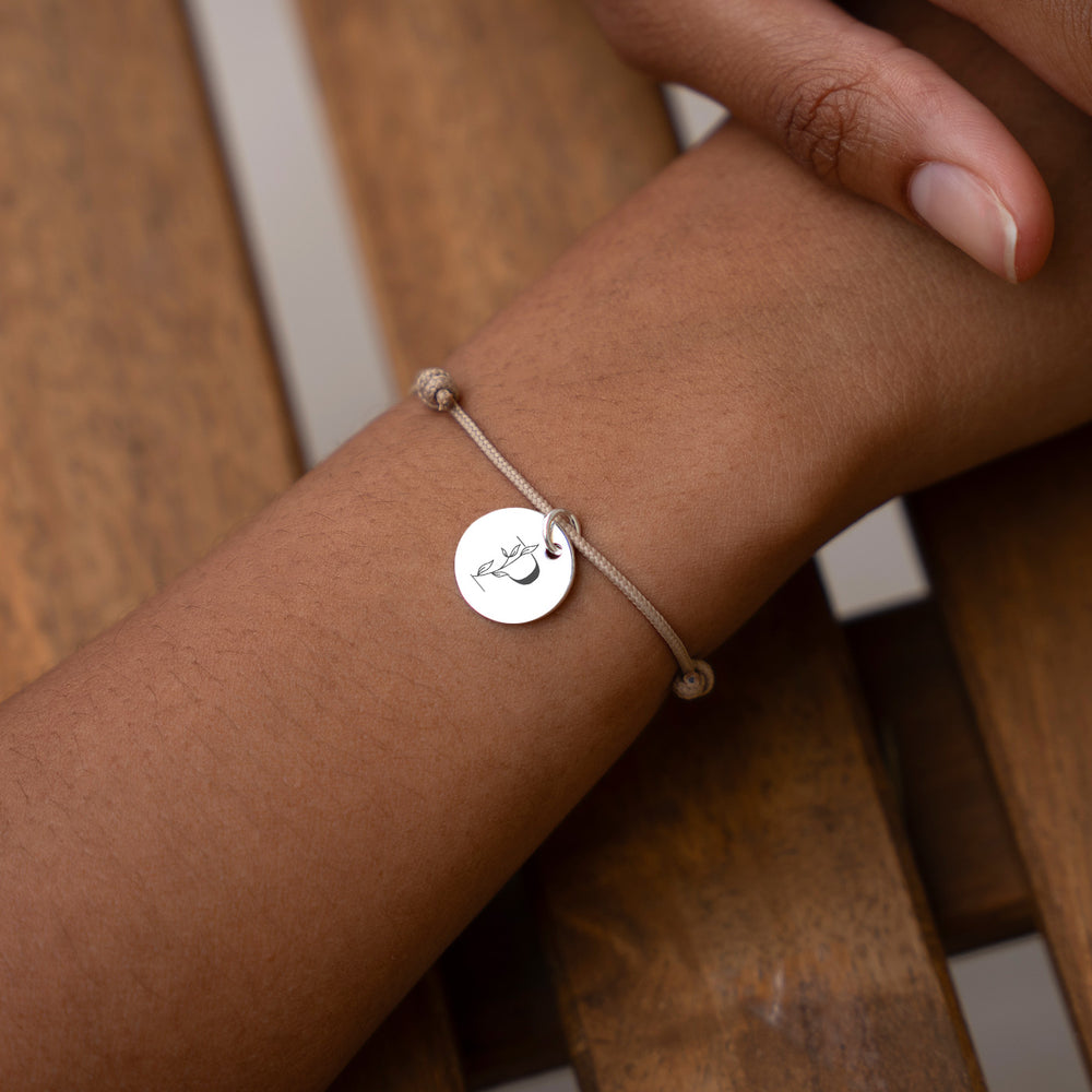 Un bracelet argenté de 15mm, orné d'une médaille personnalisée, avec un cordon beige.