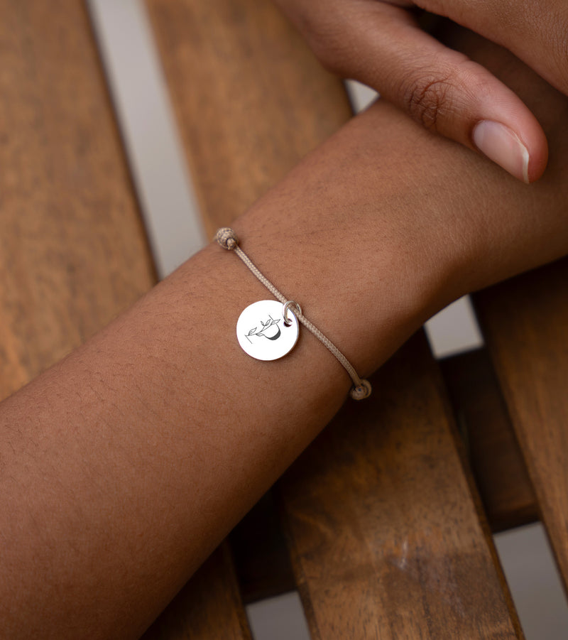 Un bracelet argenté de 15mm, orné d'une médaille personnalisée, avec un cordon beige.